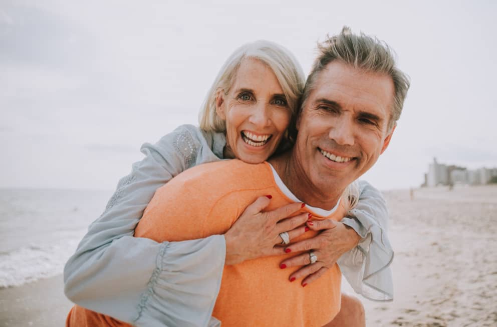 A laughing woman embraces a man from behind on the beach.
